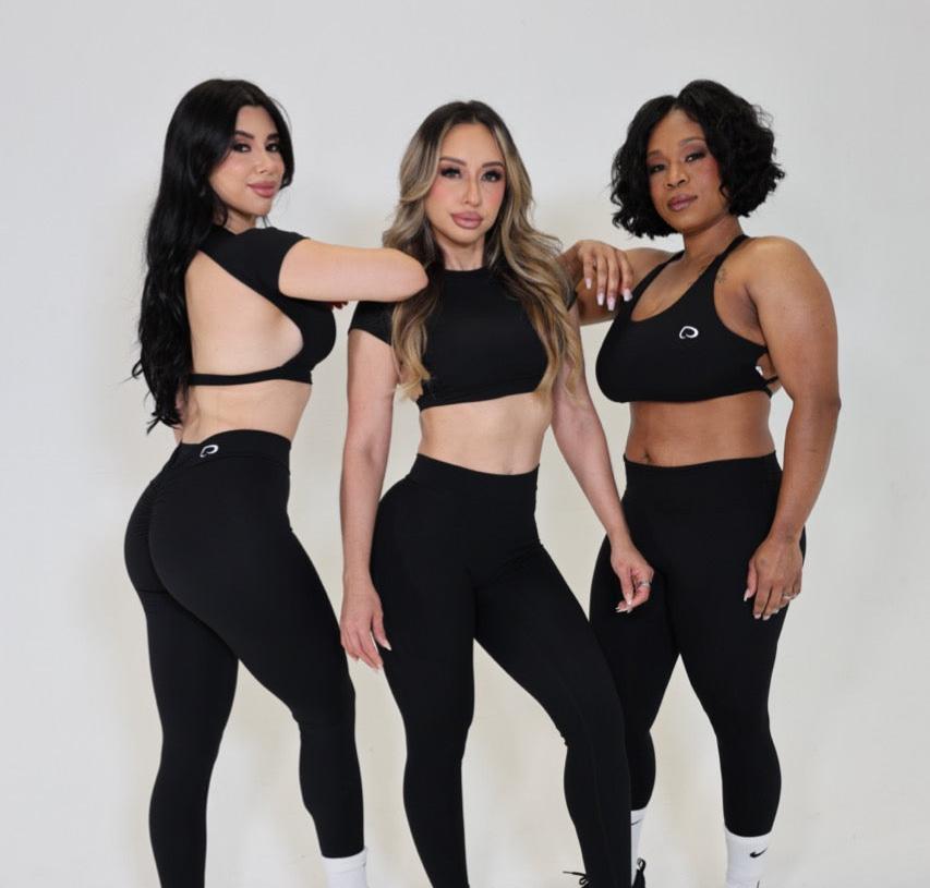 Three women standing side by side facing the camera. The first woman is wearing a black Muscle Mami cropped tee and timeless black leggings. The second woman is also wearing a black muscle mami cropped tee and black timeless leggings. The last woman is wearing double cross timeless black sports bra and timeless black leggings. 